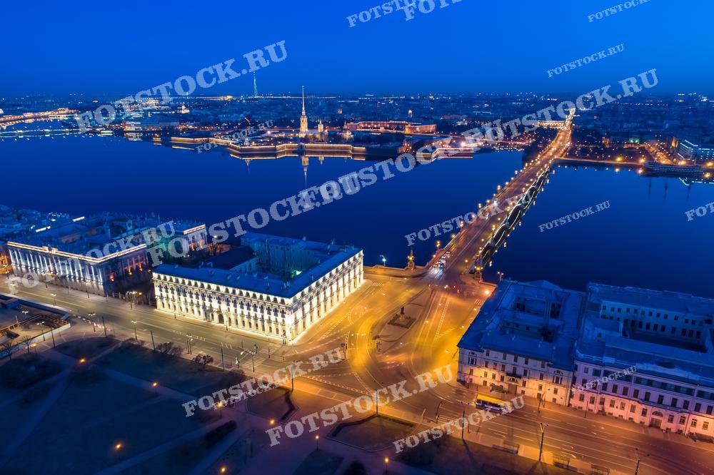 На фотографии изображен ночной вид Санкт-Петербурга, снятый с высоты. На переднем плане виден Троицкий мост через Неву, на заднем - знаменитая Петропавловская крепость. Река Нева отражает свет фонарей, создавая чарующую атмосферу. Здания на набережной также подсвечены, добавляя тепла в ночной пейзаж города.